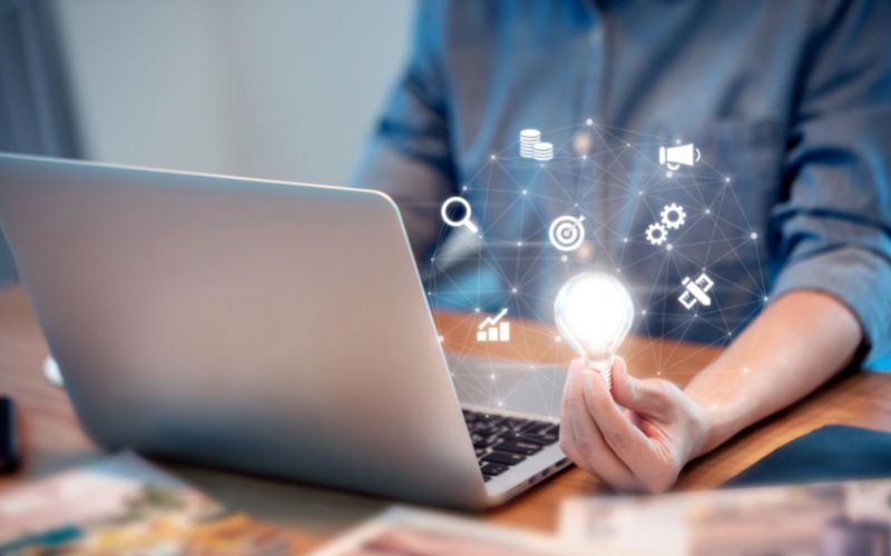 Graphics and a light bulb floating over laptop keyboard with a blurred vision of a person sitting behind it