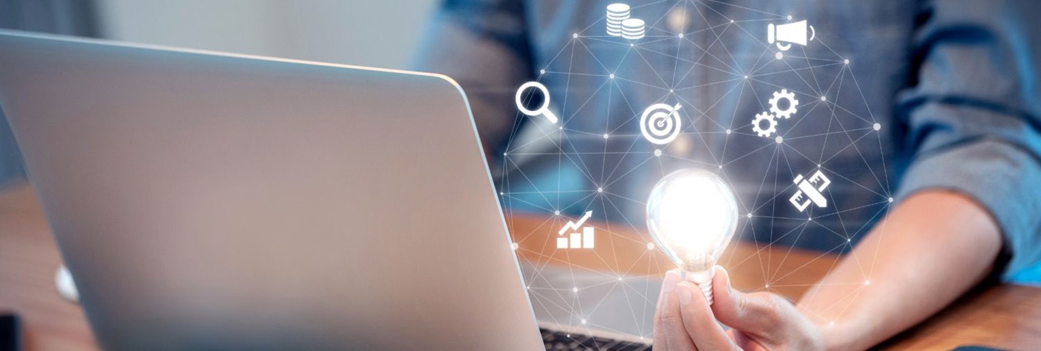 A laptop with graphics and a lightbulb floating above it with the blurred view of a person behind it